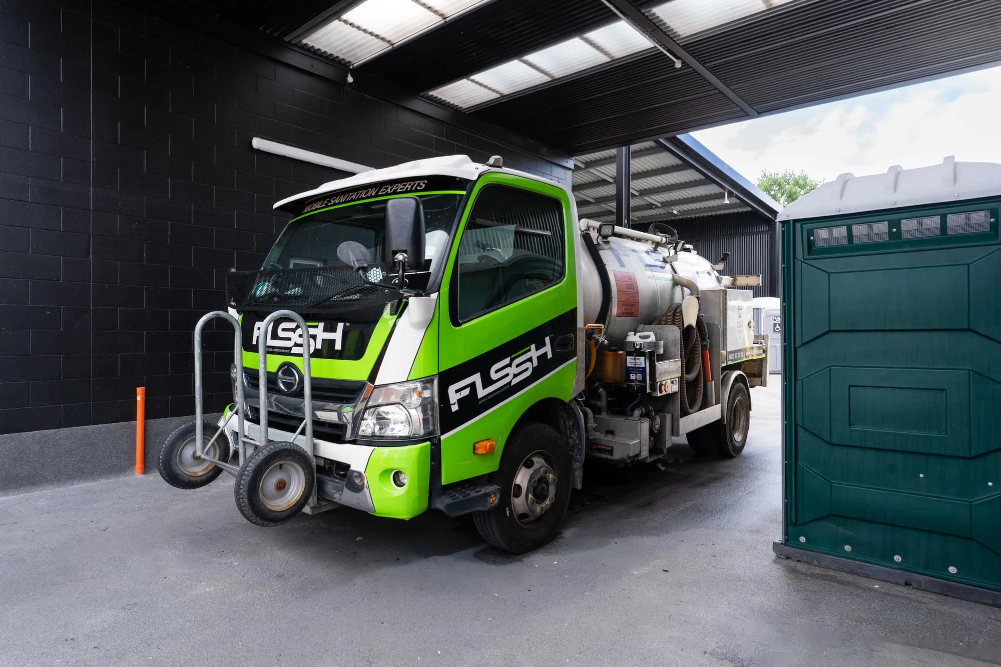 Green FLSSH service truck under the depot canopy beside a portable toilet