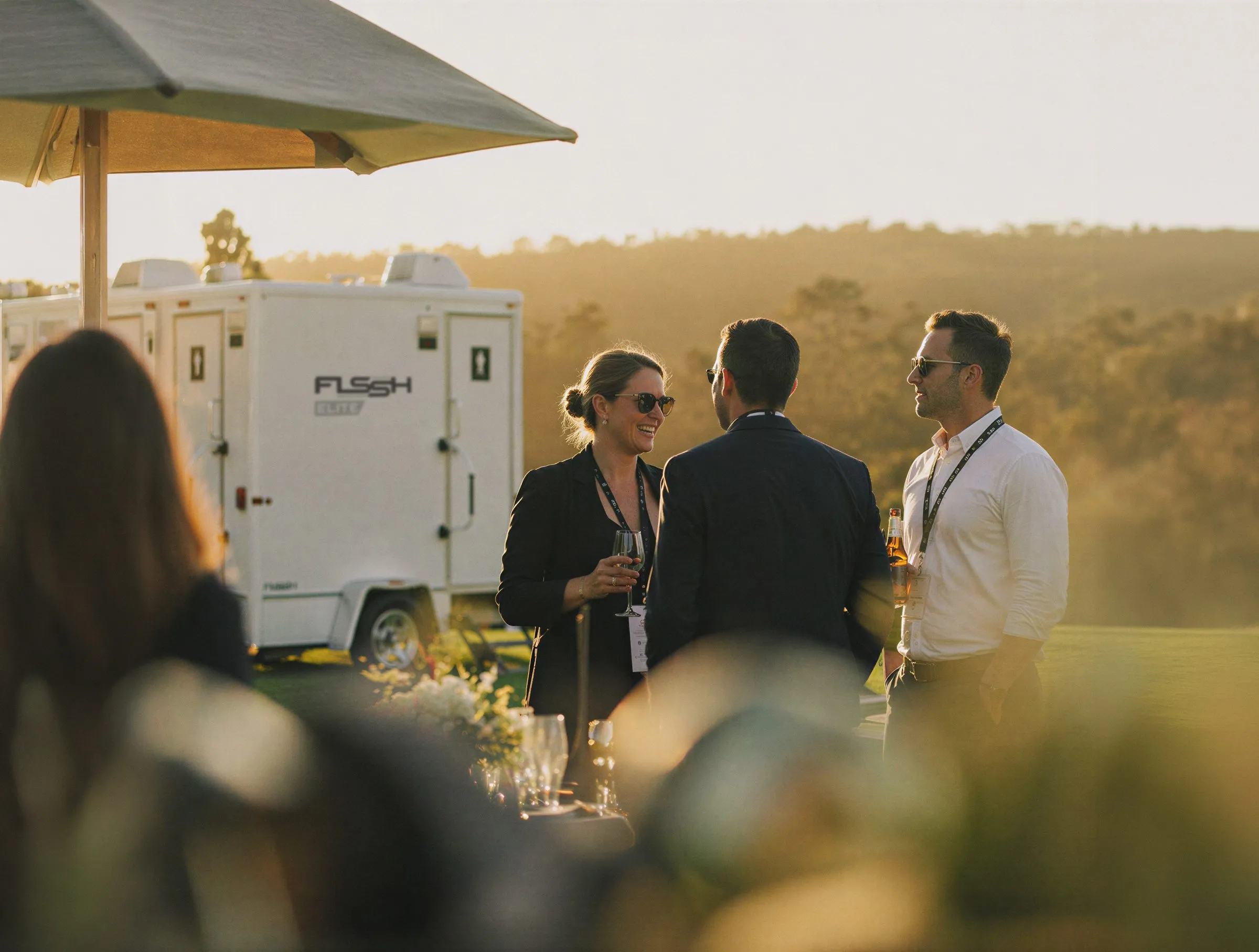 Guests talking at an outdoor corporate event with an FLSSH Elite restroom trailer behind them